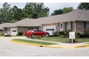 This is a brick, single-story retirement community building with parked cars and surrounding greenery.