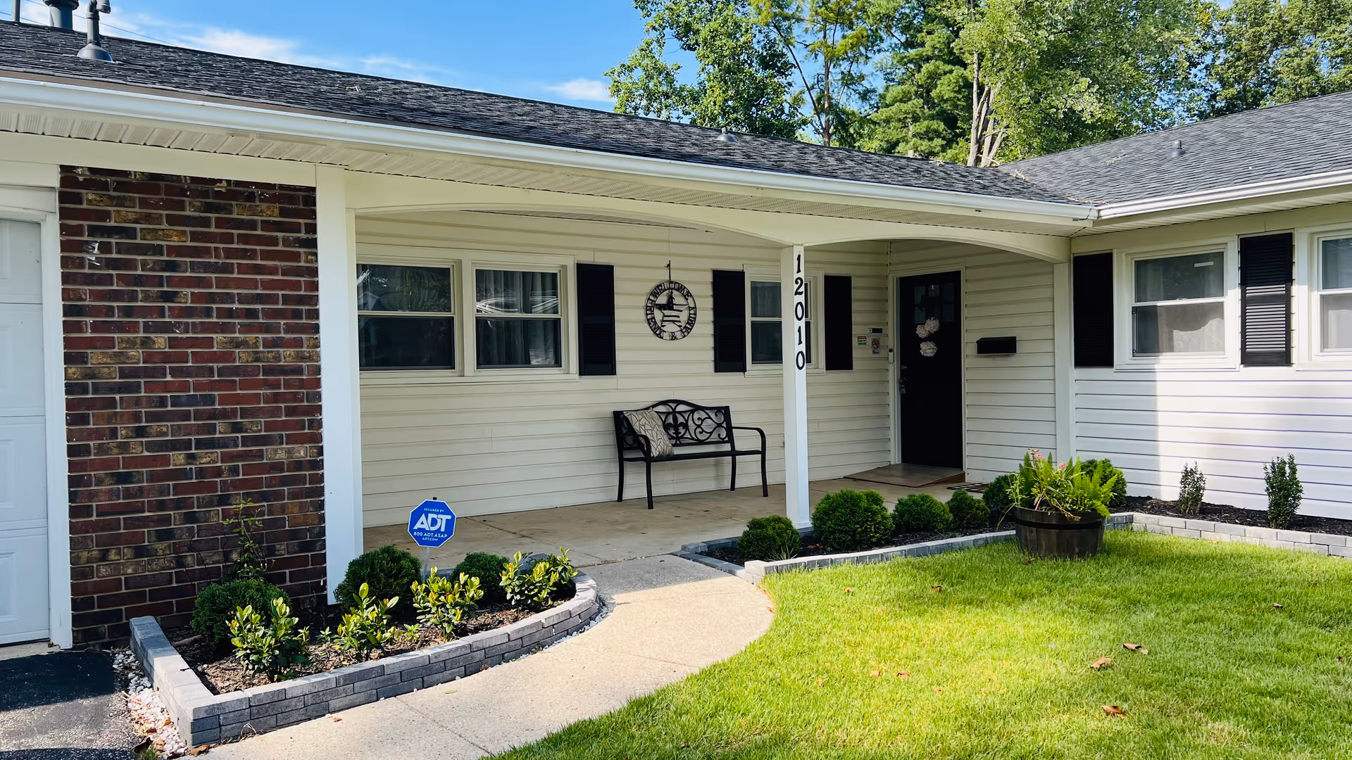 Front exterior of Asinza Assisted Living House with a porch bench, shrubs, lawn, and security sign.