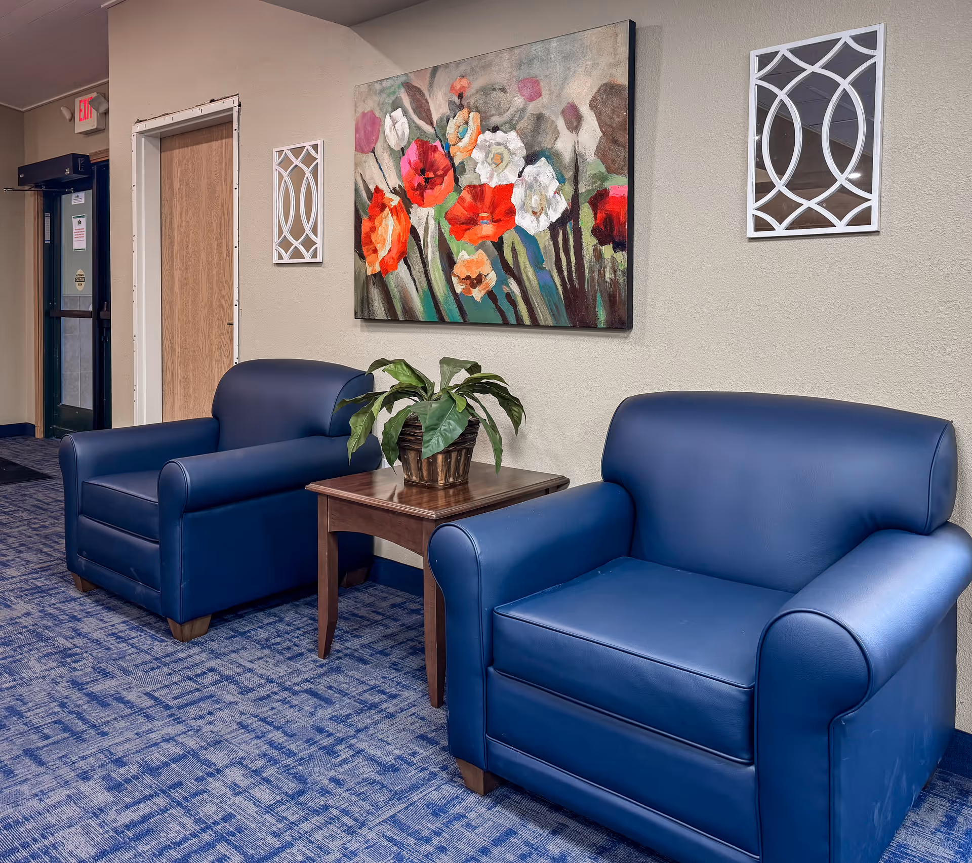 Two blue leather armchairs separated by a wooden side table with a potted plant on it, set against a beige wall with a colorful floral painting and two decorative white wall hangings. The floor is covered with a blue patterned carpet, and a door and exit sign are visible in the background.