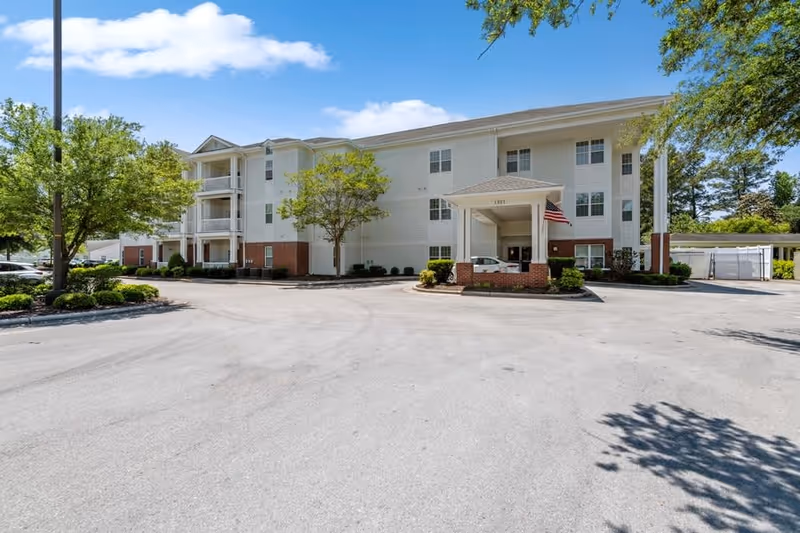 Exterior view of a three-story senior living facility building with white siding and brick accents, surrounded by trees and a paved driveway under a blue sky with some clouds. An American flag is displayed near the entrance.