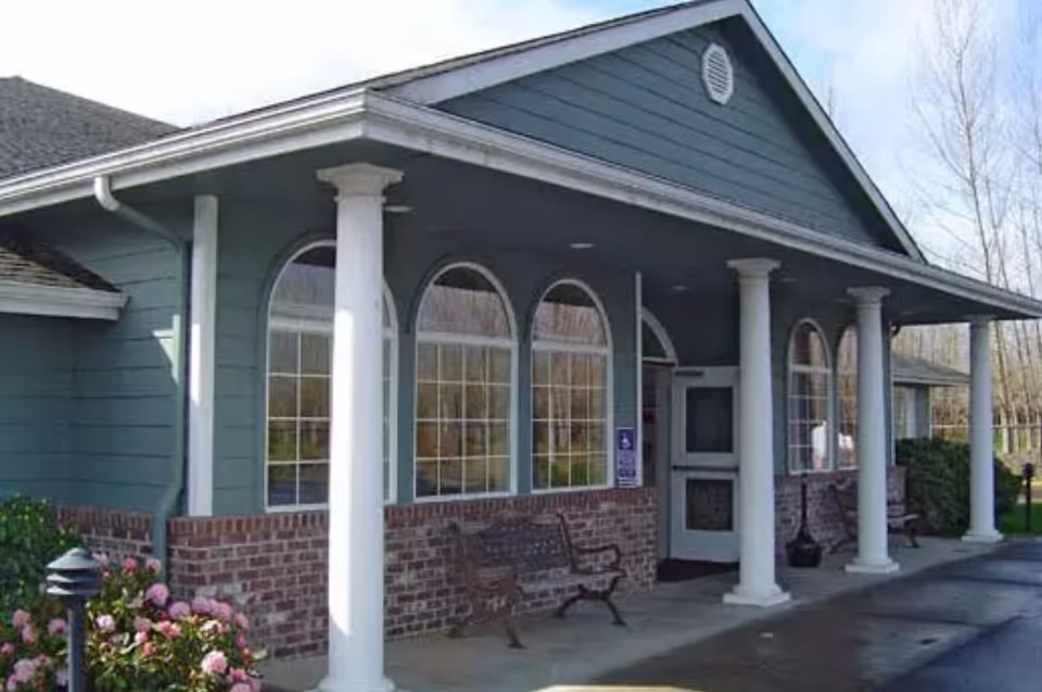 Exterior view of a building with blue siding and brick lower walls, featuring white columns supporting a covered porch area with benches and arched windows.