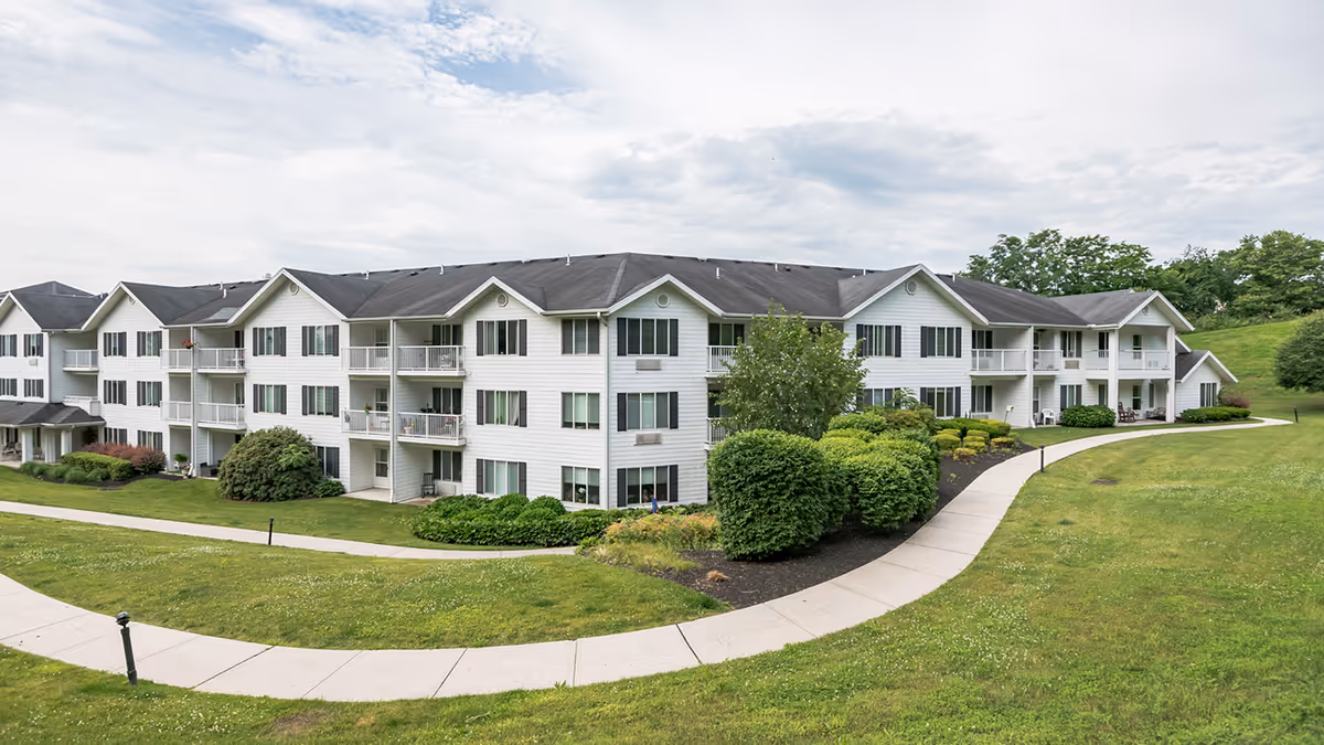 Exterior view of a large, white, three-story senior living facility building with multiple balconies and windows, surrounded by well-maintained green lawns, bushes, and a curved concrete walkway under a partly cloudy sky.