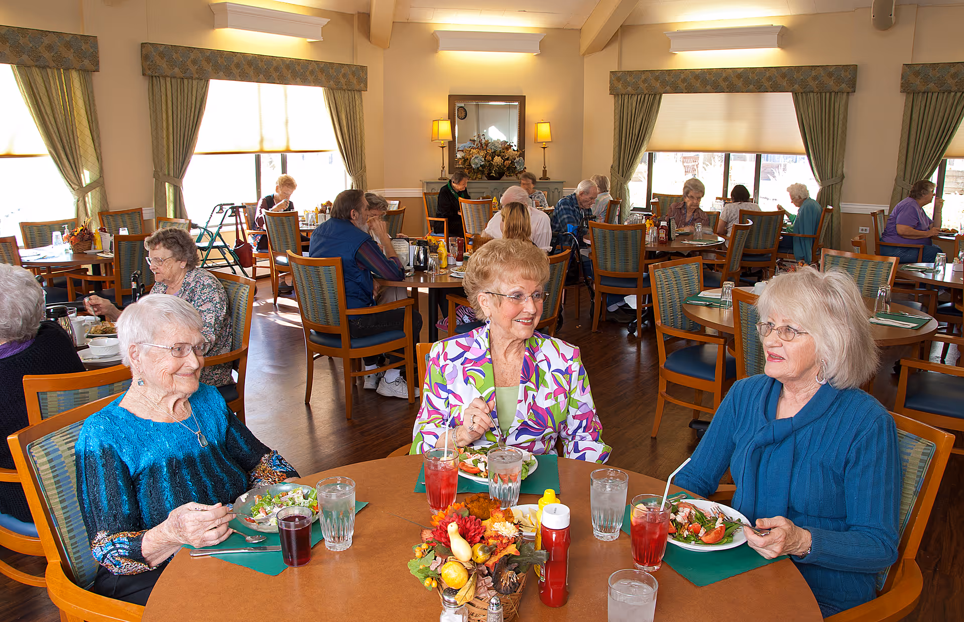 A dining room in Eaton Senior Communities with elderly residents seated at tables enjoying their meals. The room is well-lit with large windows covered by curtains, and the tables are set with plates of food, drinks, and condiments. Three elderly women are seated at the foreground table, smiling and conversing while eating salad.