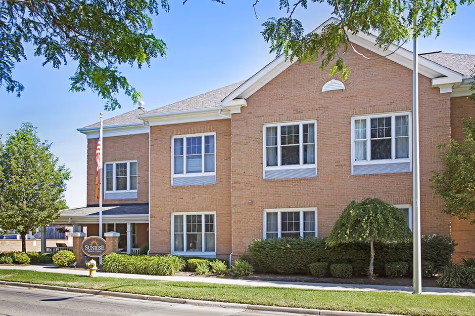 Exterior view of a two-story brick building with multiple windows, surrounded by green shrubs and trees under a clear blue sky. A sign near the entrance reads 'Sunrise on Vernier'.