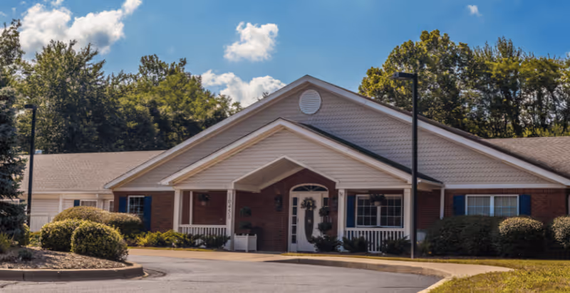 Single-story brick senior living building with a covered front entrance, driveway, and landscaped shrubs under a blue sky.