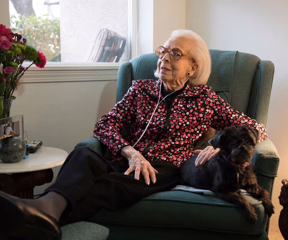 An elderly woman sits in a teal armchair with a small dog beside her.