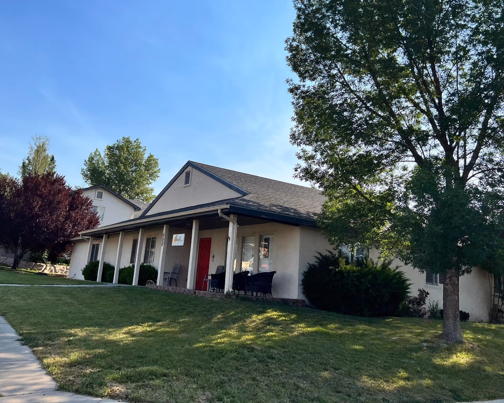 Exterior view of a single-story building with a covered porch, red front door, and several chairs on the porch. The building is surrounded by green grass, trees, and bushes under a clear blue sky.