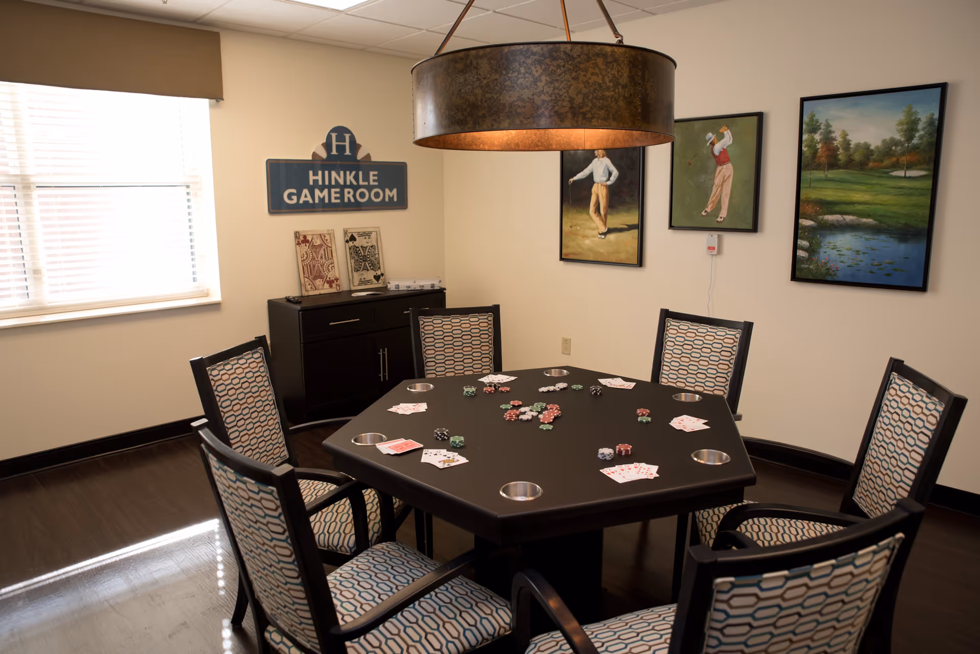 A game room with a hexagonal poker table surrounded by six patterned chairs. The table has playing cards and poker chips scattered on it. On the wall, there is a sign that reads 'Hinkle Gameroom' and three framed paintings depicting golf scenes. A large circular pendant light hangs above the table, and a window with blinds lets in natural light.