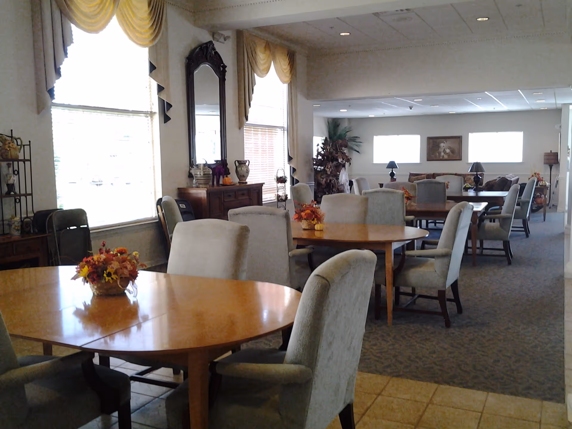 A bright dining room with round wooden tables, upholstered chairs, floral centerpieces, and large windows in a senior living common area.