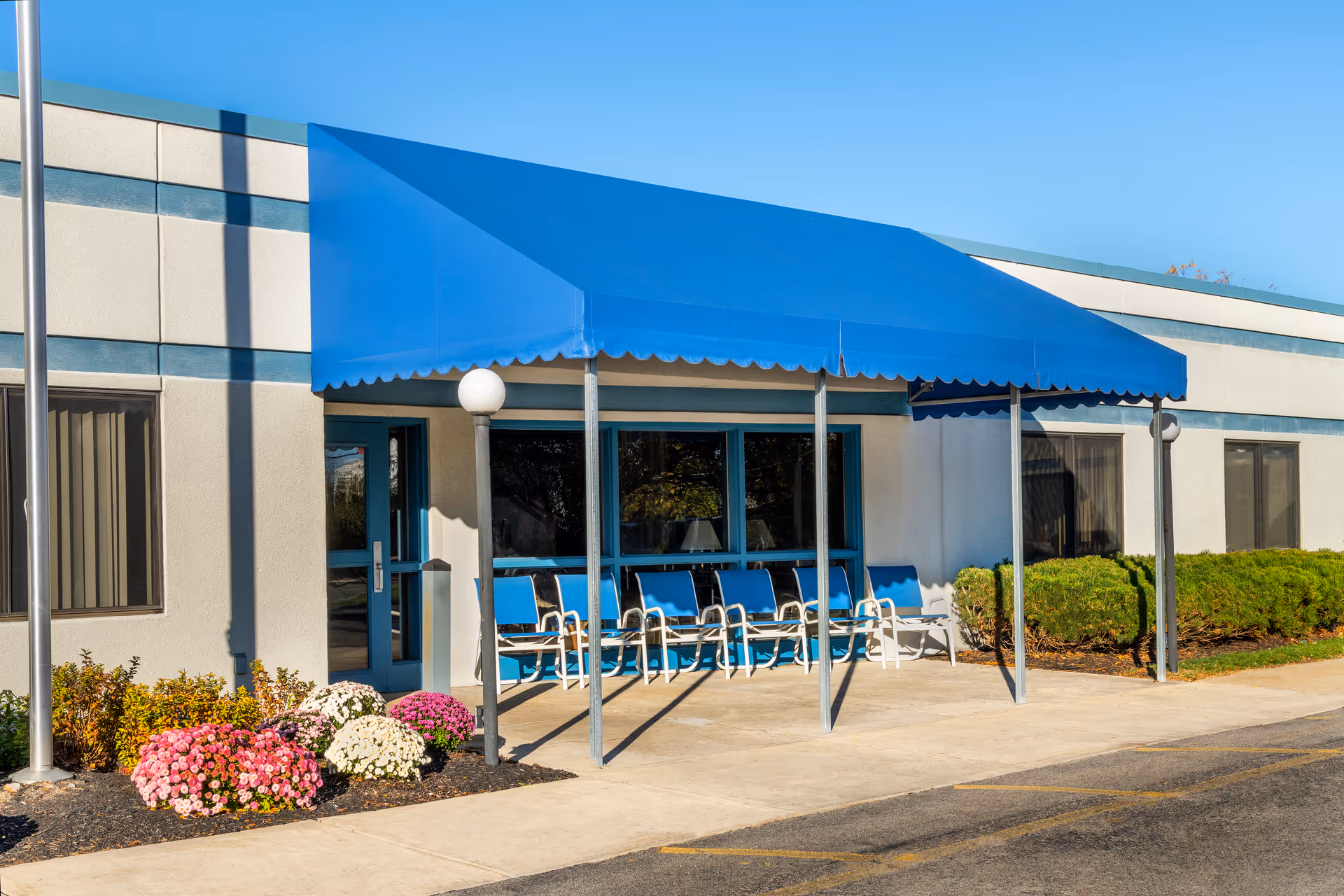Exterior view of a building entrance with a blue awning and several blue chairs lined up underneath it. There are windows and a door behind the chairs, with some bushes and colorful flowers planted along the building. The sky is clear and blue.