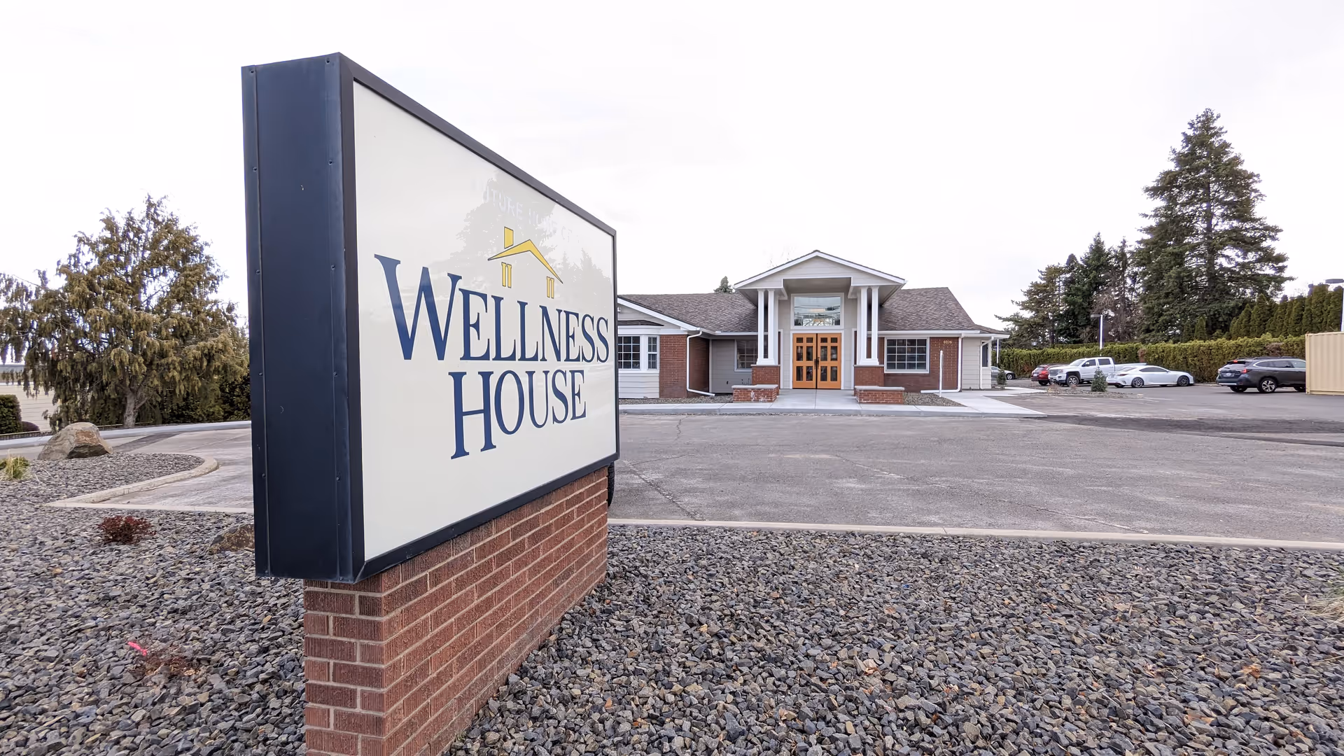 Exterior view of a Wellness House facility entrance with a sign and parking lot.