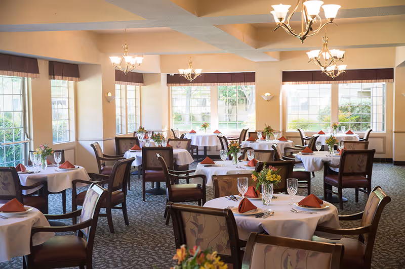 A bright and elegant dining room with multiple round tables covered with white tablecloths, each set with glassware, silverware, and red folded napkins. The room has large windows letting in natural light, floral centerpieces on each table, patterned carpet, and chandeliers hanging from the ceiling.