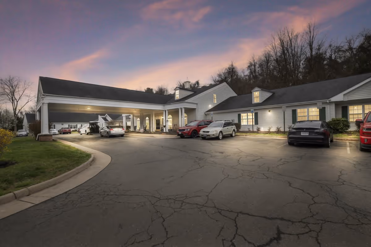 Exterior view of a senior living facility named Spring Oak at Warrenton during dusk, showing a driveway with several parked cars and a covered entrance with columns. The building has white siding, multiple windows with lights on, and a backdrop of trees under a purple and pink sky.