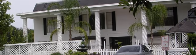 White two-story retirement facility with columns, palm trees, a fenced front yard, and a parked car.