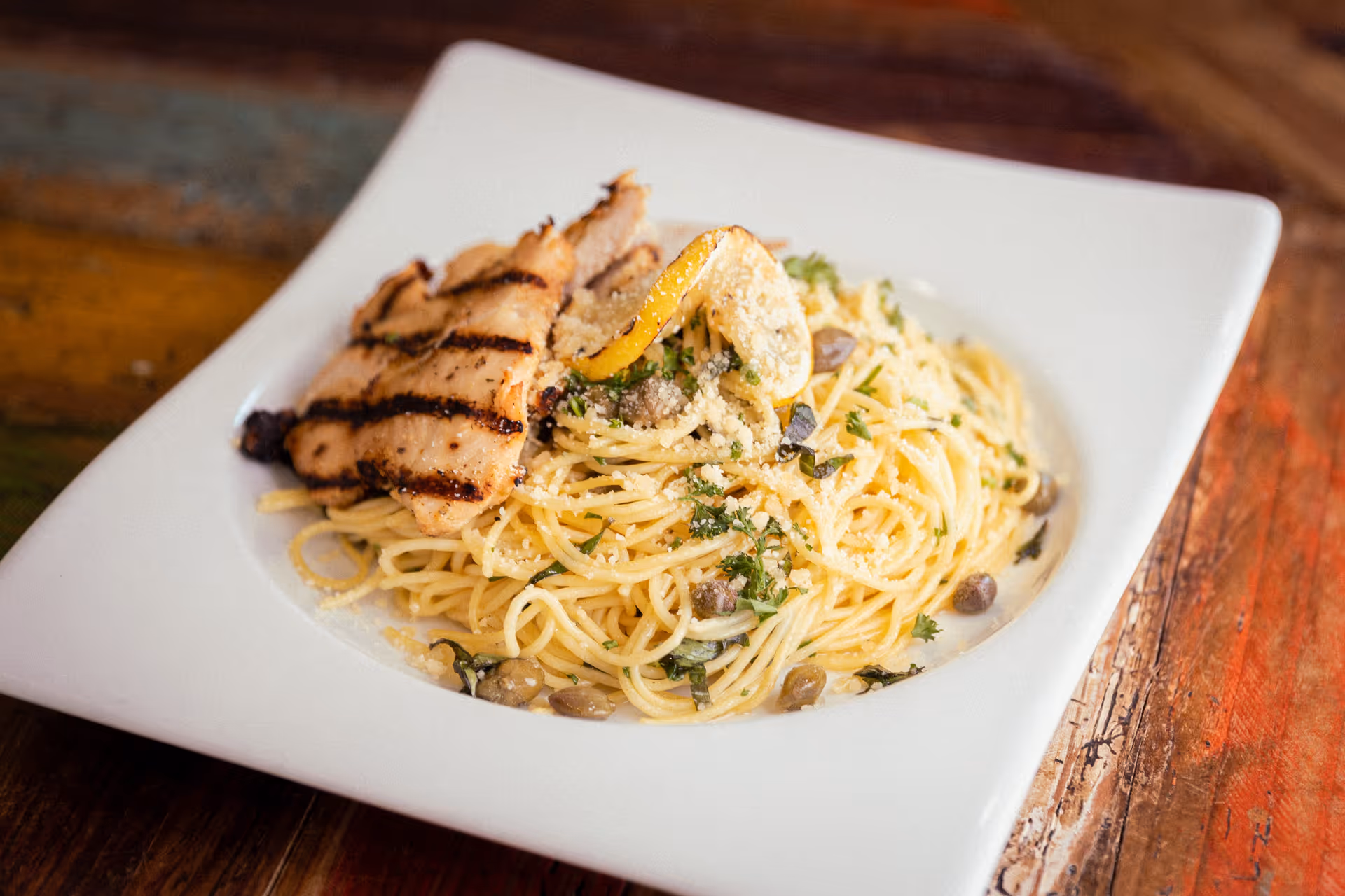A white square plate with a serving of spaghetti pasta garnished with herbs, capers, grated cheese, and a grilled lemon slice, topped with two pieces of grilled chicken breast with grill marks, placed on a rustic wooden table.