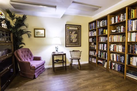 A cozy reading room with tall bookshelves, a purple armchair, a table lamp, and framed art.