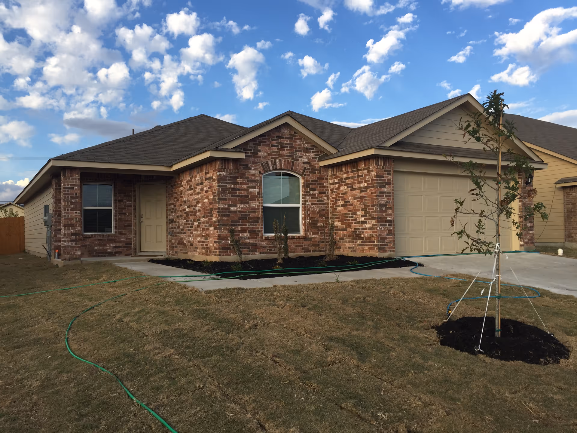 Single-story brick house with attached garage, front yard, and a young tree under a blue sky.