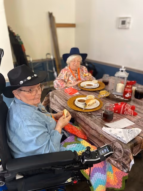 Two elderly women wearing hats sit at a decorated dining table with plates of food in a communal room.
