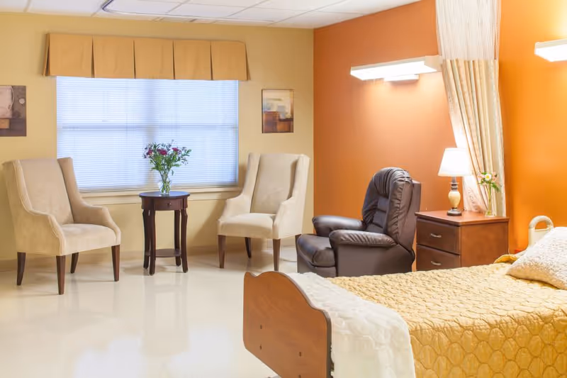 A cozy room with a single bed covered in a yellow quilt, a wooden nightstand with a lamp and a small vase of flowers, a brown leather recliner chair, two beige upholstered armchairs, and a small round wooden table with a vase of flowers in front of a window with blinds and a yellow valance.
