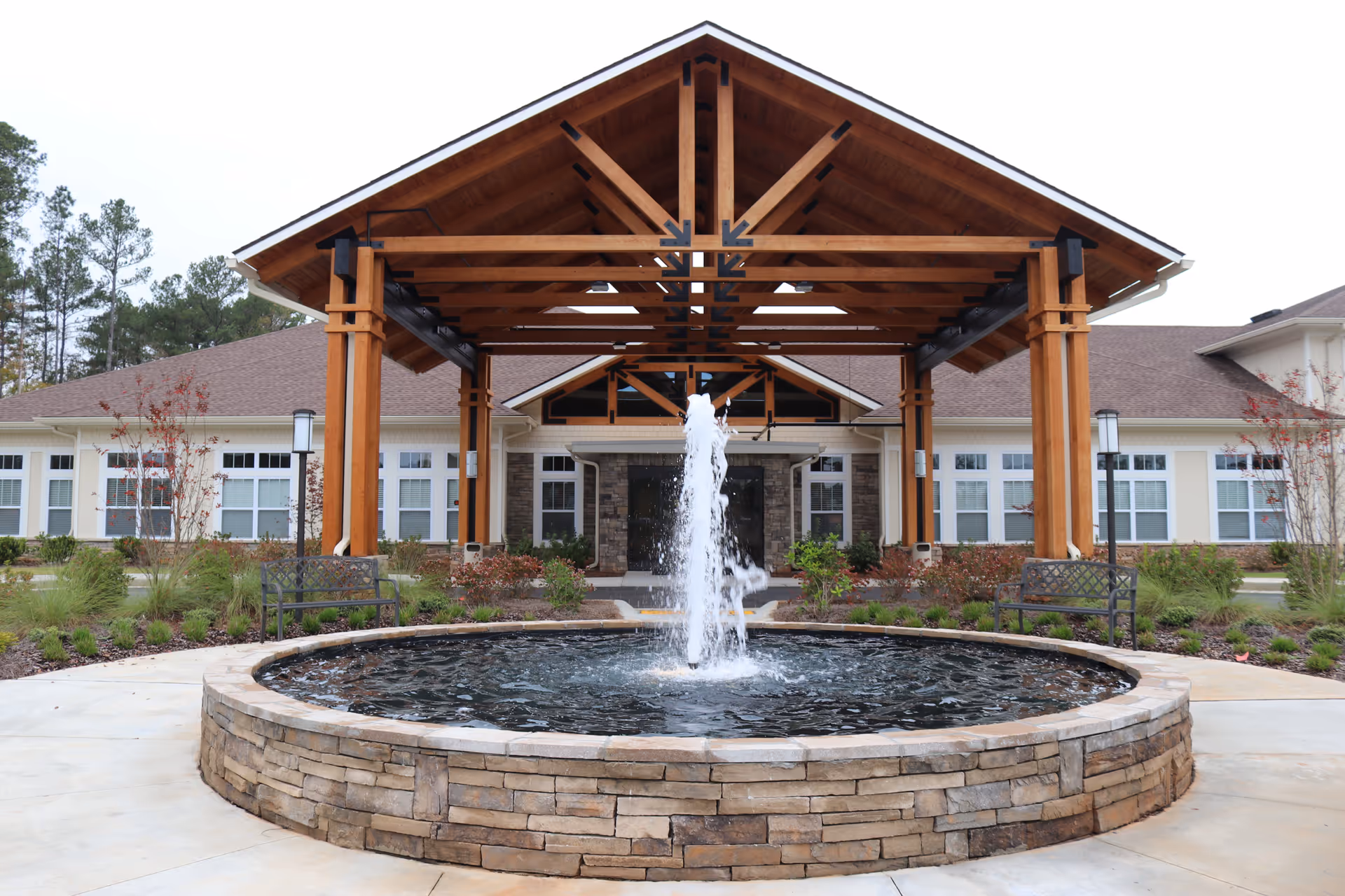 Front exterior view of a senior living facility named Prema at Ashton Hills featuring a large wooden pergola structure over the entrance, a circular stone fountain with water spraying upwards in the foreground, benches on either side, and landscaped greenery around the building.