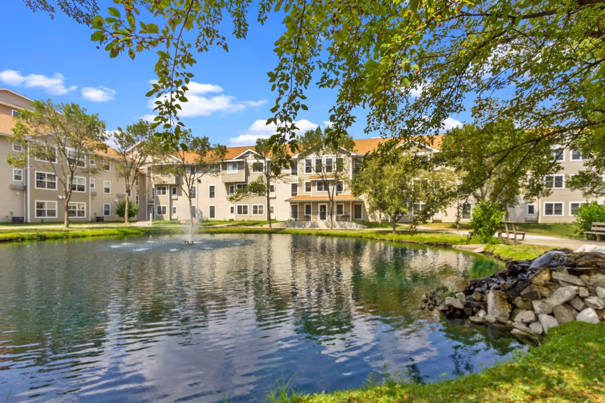 Exterior view of a multi-story senior living building beside a pond with a fountain and surrounding trees.