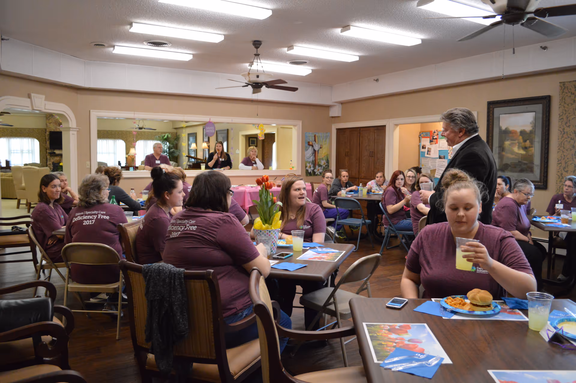 A group of people gathered in a dining room setting, many wearing matching maroon shirts with 'Deficiency Free 2017' printed on the back. They are seated at tables with food and drinks, engaging in conversation. The room has warm lighting, ceiling fans, and a large mirror on one wall reflecting part of the scene.