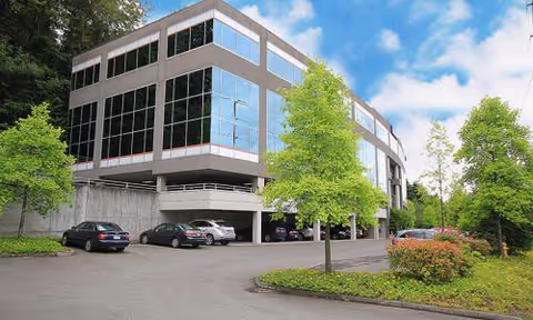 A modern, curved building with large glass windows, a parking area, and surrounding trees.