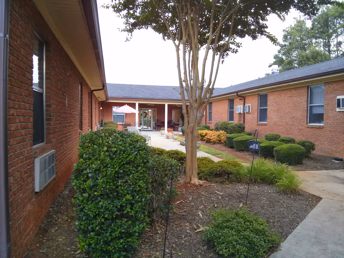 Brick-walled courtyard with a central tree, trimmed shrubs and a walkway leading to outdoor seating and an umbrella.
