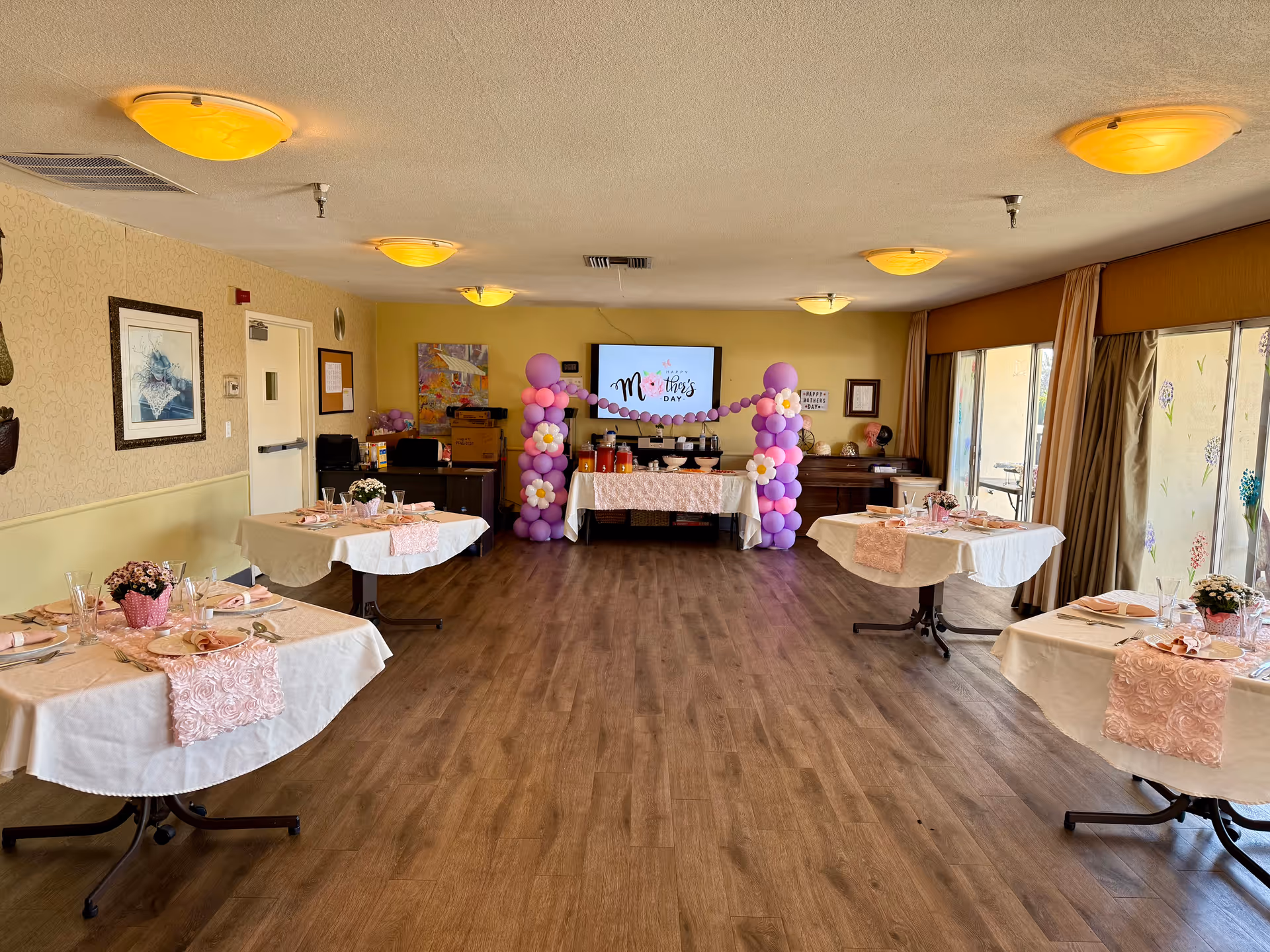 A decorated senior living dining room with pink party table settings and balloon arches.