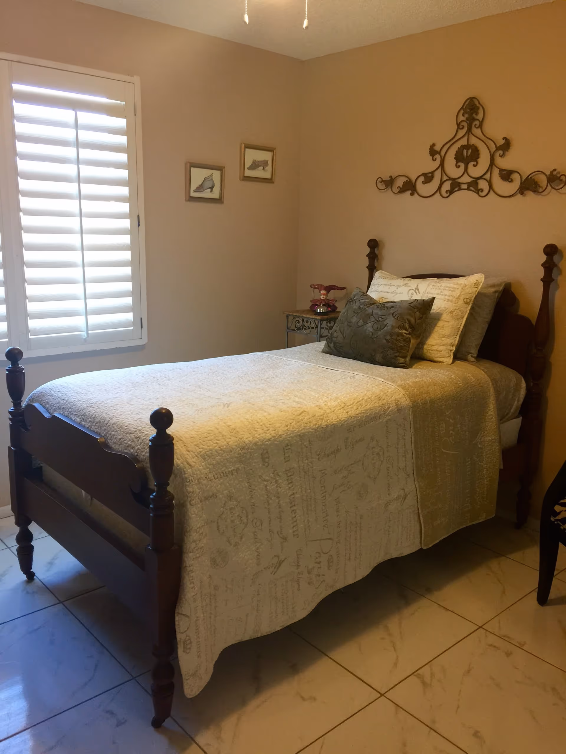 A small bedroom with a single wooden bed covered with a beige quilt and multiple pillows. The room has beige walls, a window with white shutters, two framed pictures of shoes on the wall, a decorative metal wall hanging above the bed, and a small side table with a pink decorative item.