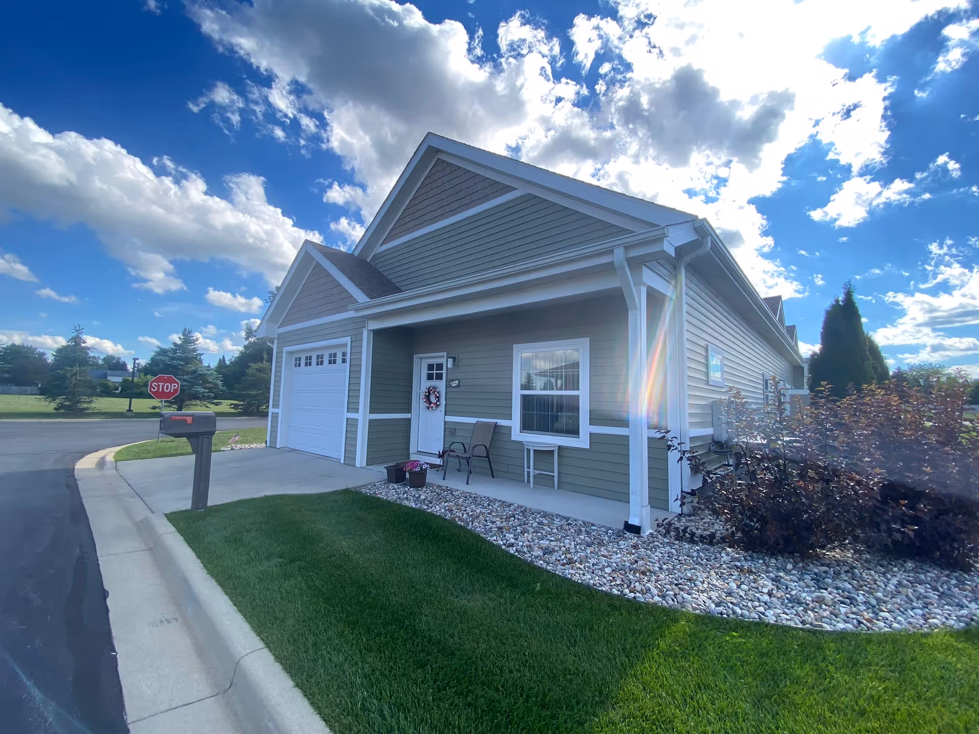 Exterior view of a single-story residential building with green siding and white trim under a partly cloudy blue sky. The building features a garage door, a white front door with a decorative wreath, a window, two chairs, a small table, and landscaping with grass, rocks, and bushes. A mailbox and a stop sign are visible near the street curb.