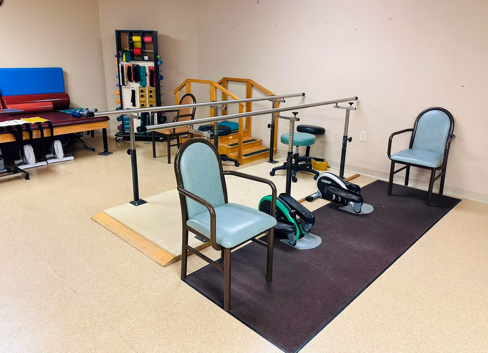Rehabilitation gym with parallel bars, exercise equipment, and chairs in a senior living facility.