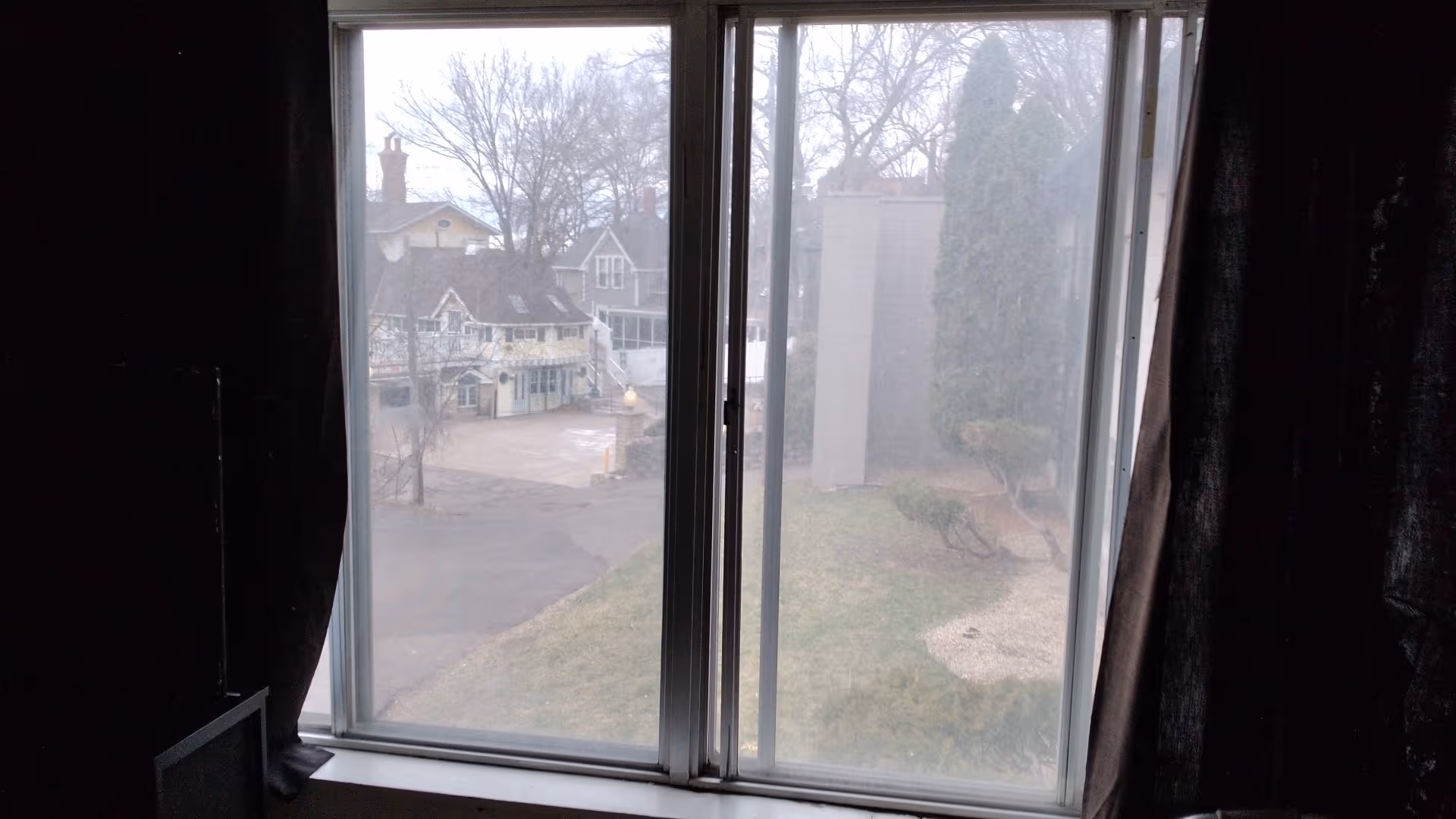 Interior view of a dark room with a large window and heavy curtains overlooking residential street.