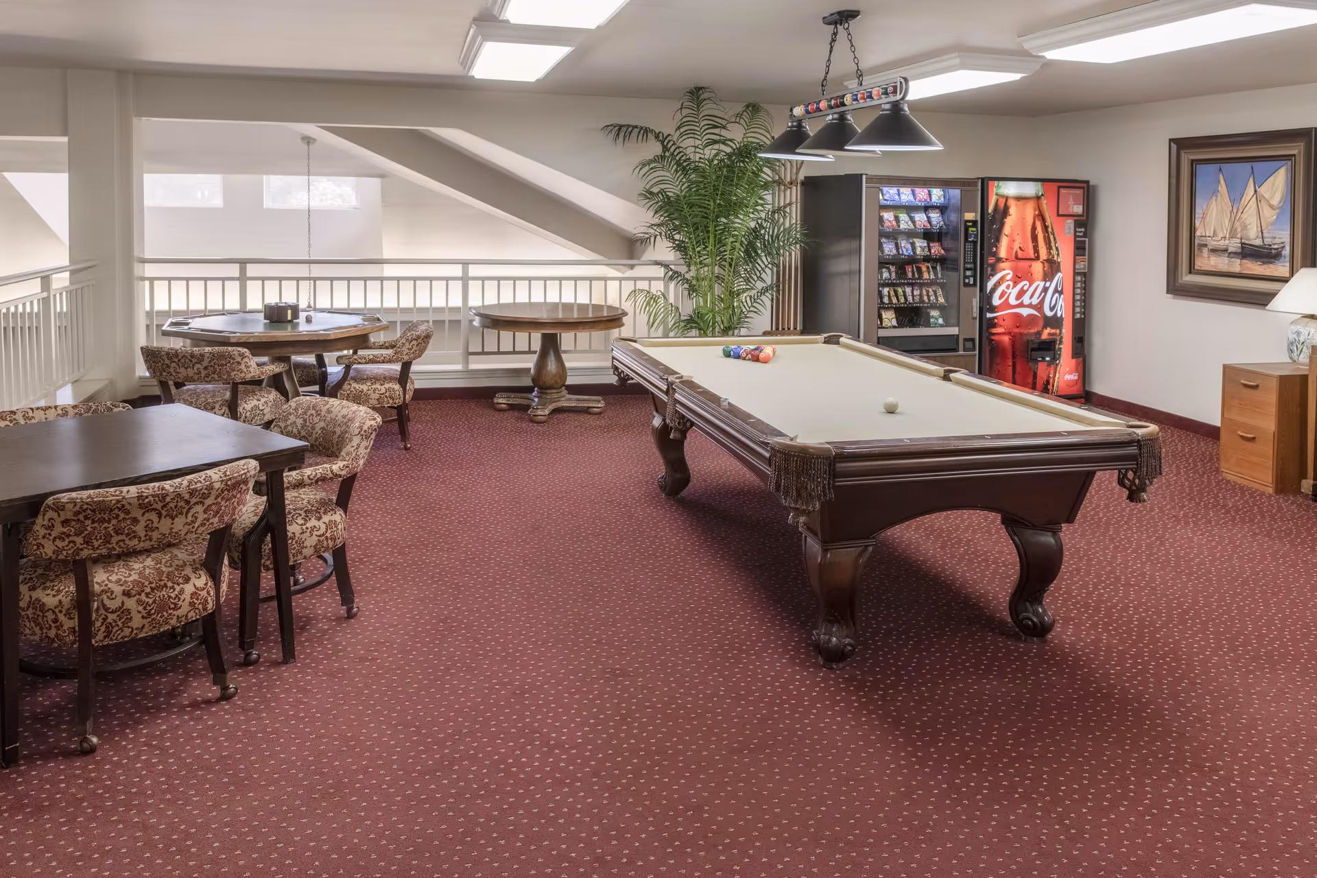 A recreational room with a pool table in the center, a vending machine with snacks and Coca-Cola drinks, several tables with patterned upholstered chairs, a large potted plant, and a framed painting of sailboats on the wall.