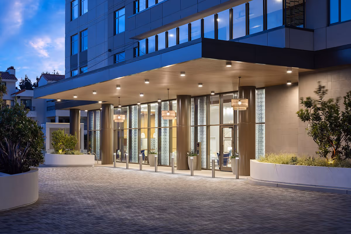 Evening view of the entrance to Belmont Village Senior Living La Jolla, featuring a modern building facade with large glass windows, illuminated hanging lights, and landscaped planters on either side of the paved driveway.