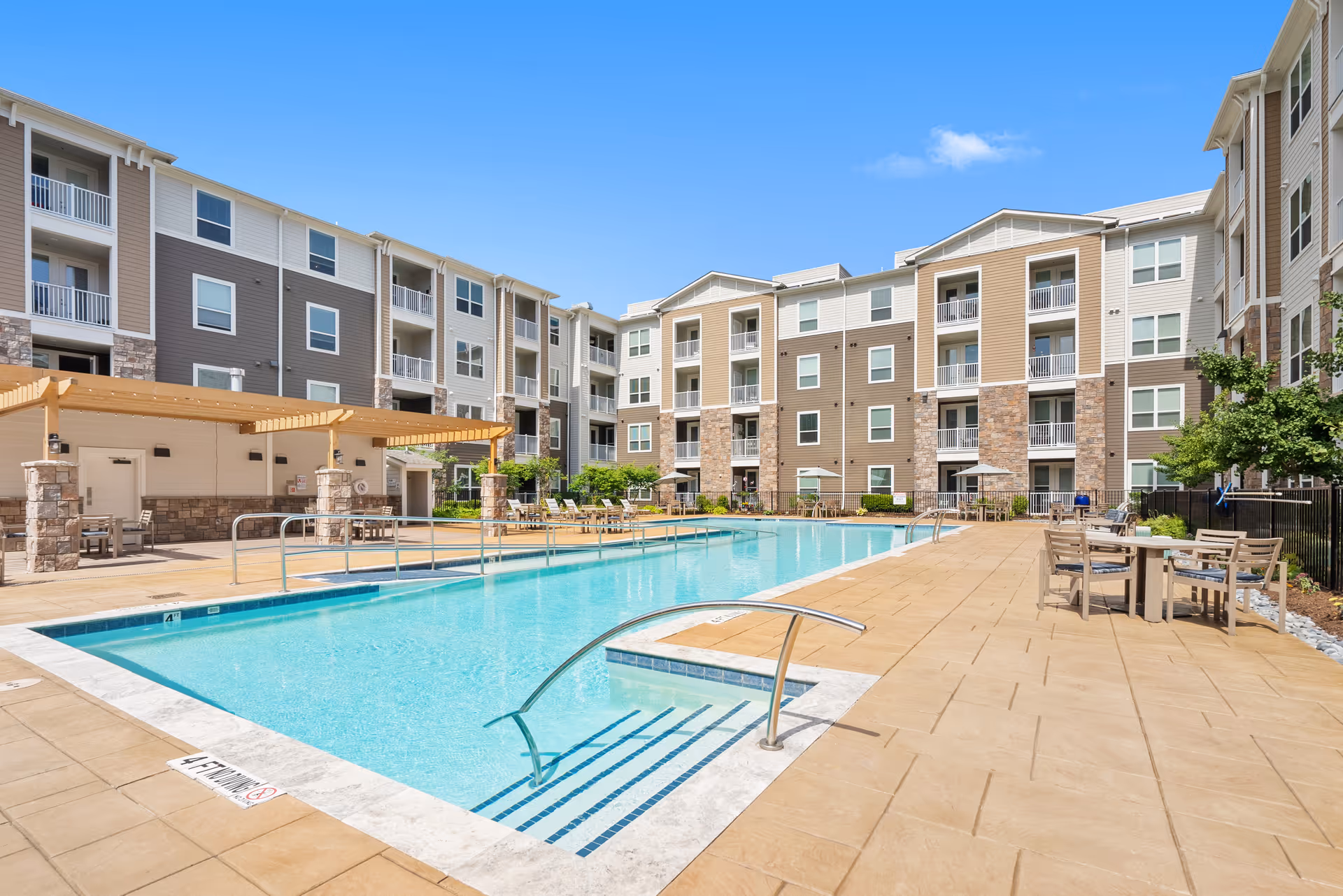 Outdoor swimming pool area at a senior living facility with lounge chairs, tables with chairs, and a pergola. The pool is surrounded by a multi-story residential building under a clear blue sky.