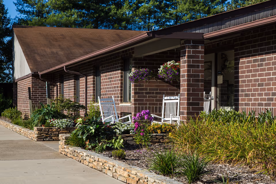 Exterior view of a brick building with a brown roof, featuring a small porch area with white rocking chairs and hanging flower baskets. The building is surrounded by landscaped flower beds with various plants and flowers, and a concrete walkway runs alongside it.