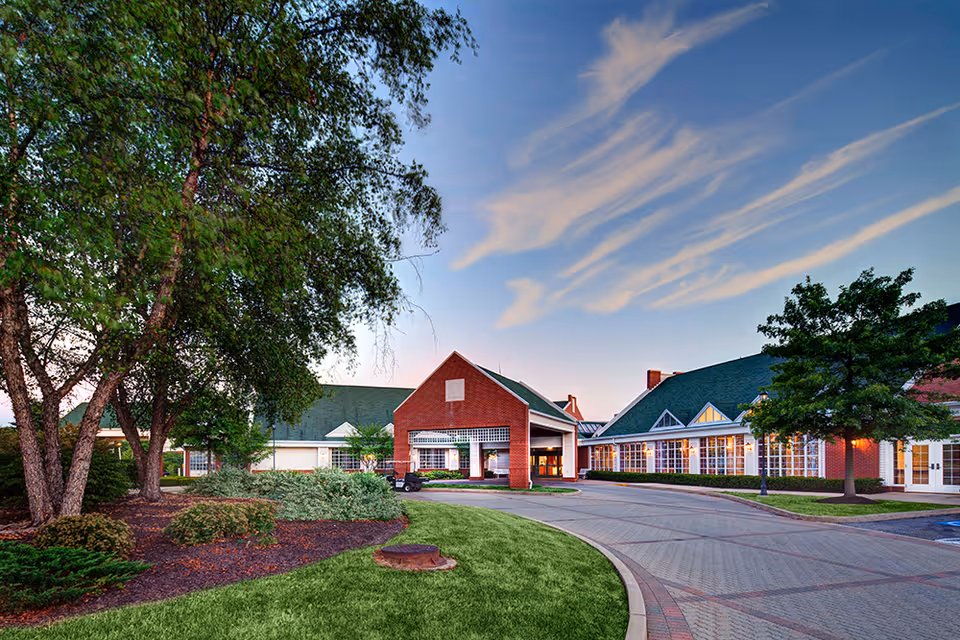 Front entrance of a red-brick retirement community building with a circular driveway, landscaped lawn and trees under a blue sky.