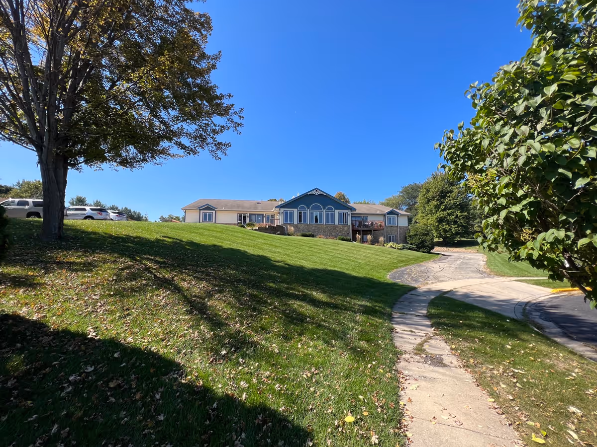 A single-story building with a blue and beige exterior situated on a grassy hill under a clear blue sky. There is a curved sidewalk and driveway leading up to the building, with trees and parked cars visible on the left side.