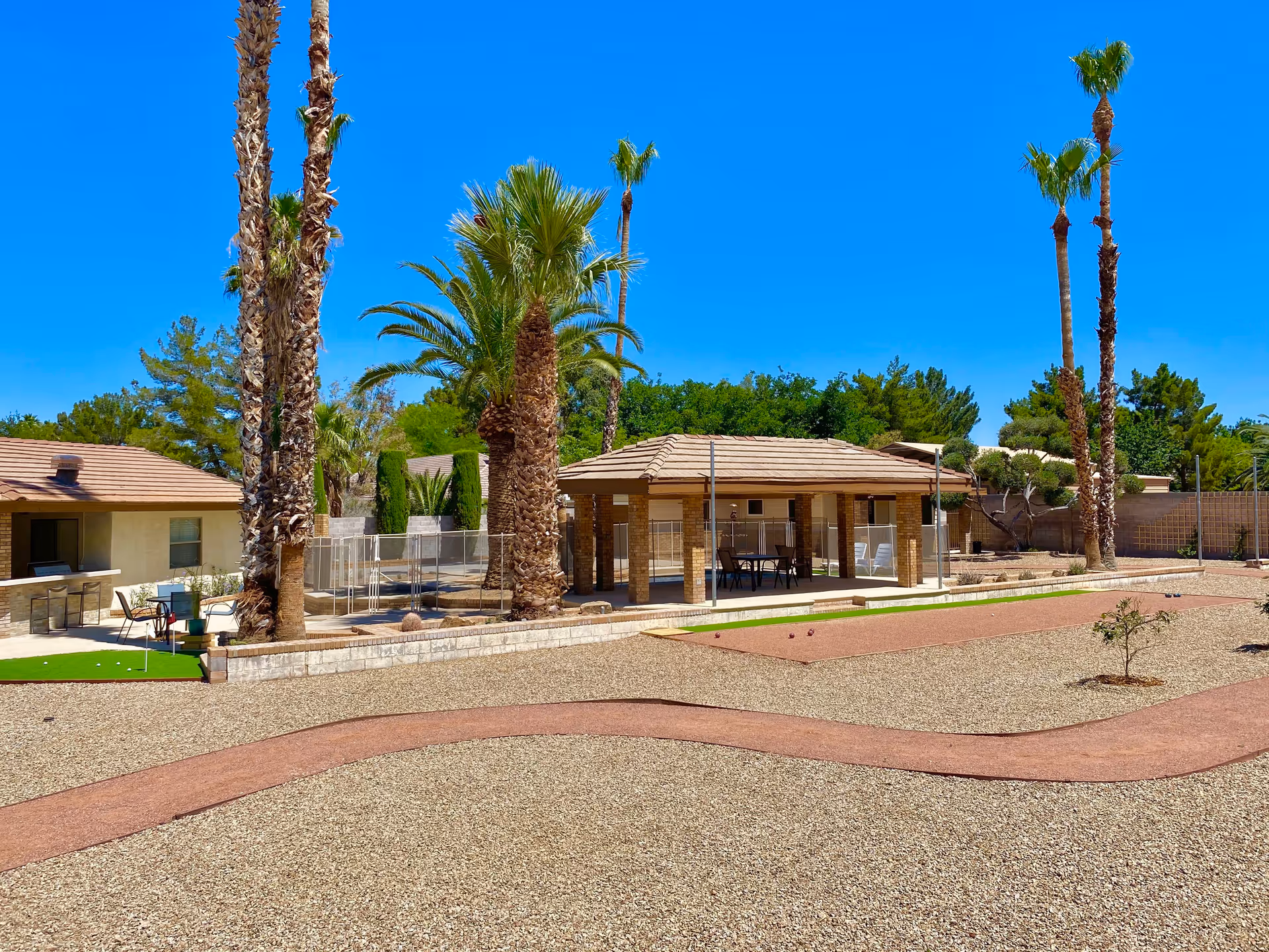 Outdoor area of The Quail House featuring a covered patio with seating, surrounded by palm trees and other greenery under a clear blue sky. The ground is covered with gravel and there is a bocce ball court in the foreground.