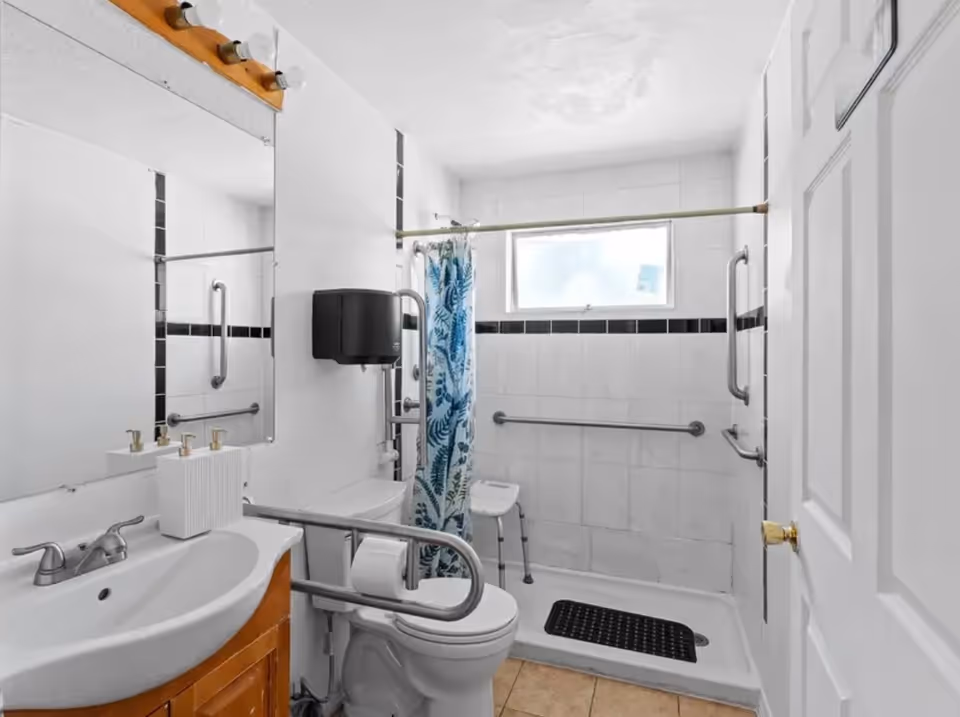 Accessible bathroom with grab bars, shower seat, sink, mirror, and toilet.