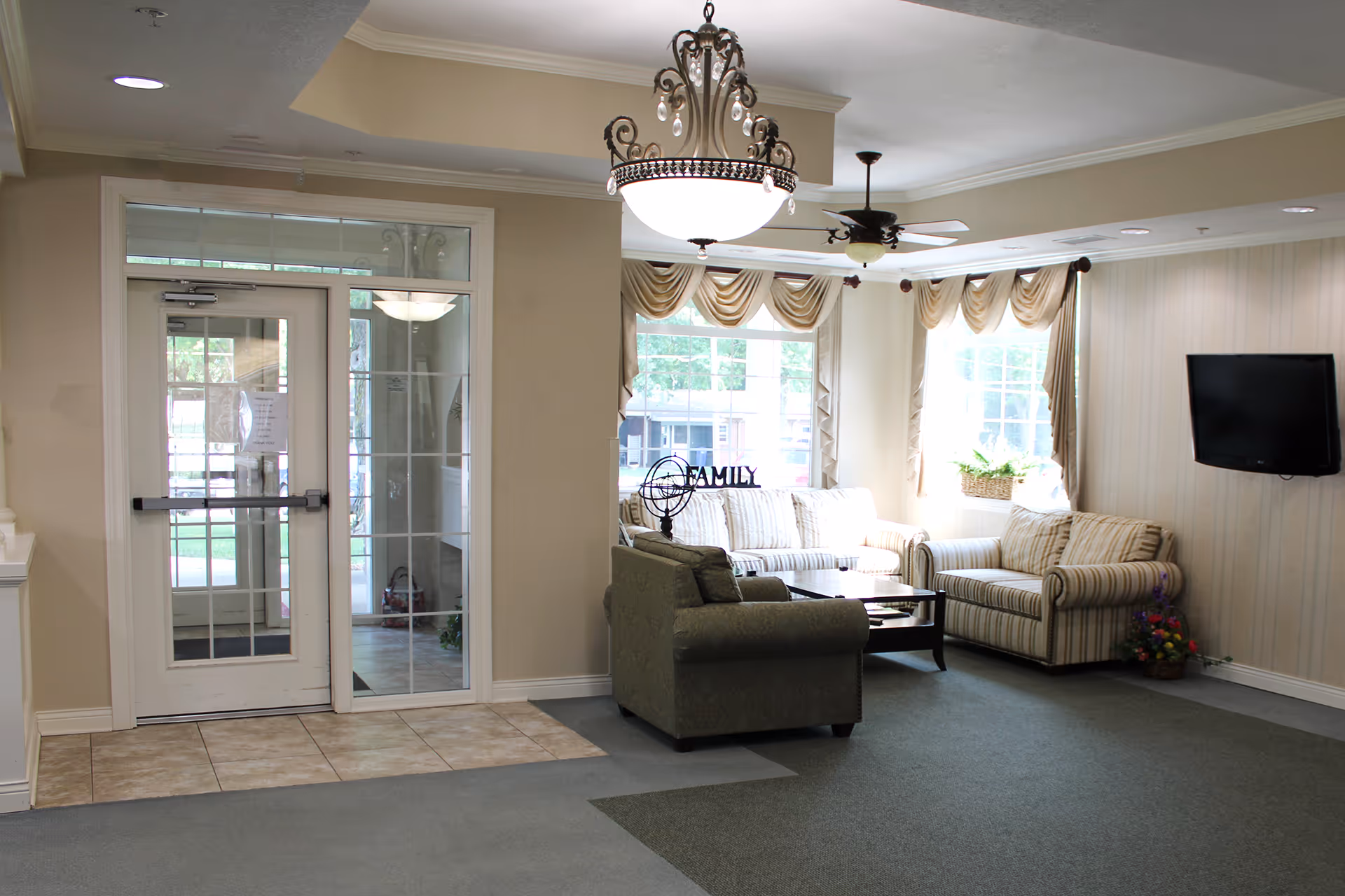 Cozy senior living lounge with beige walls, striped sofa, armchairs, coffee table, and windows.