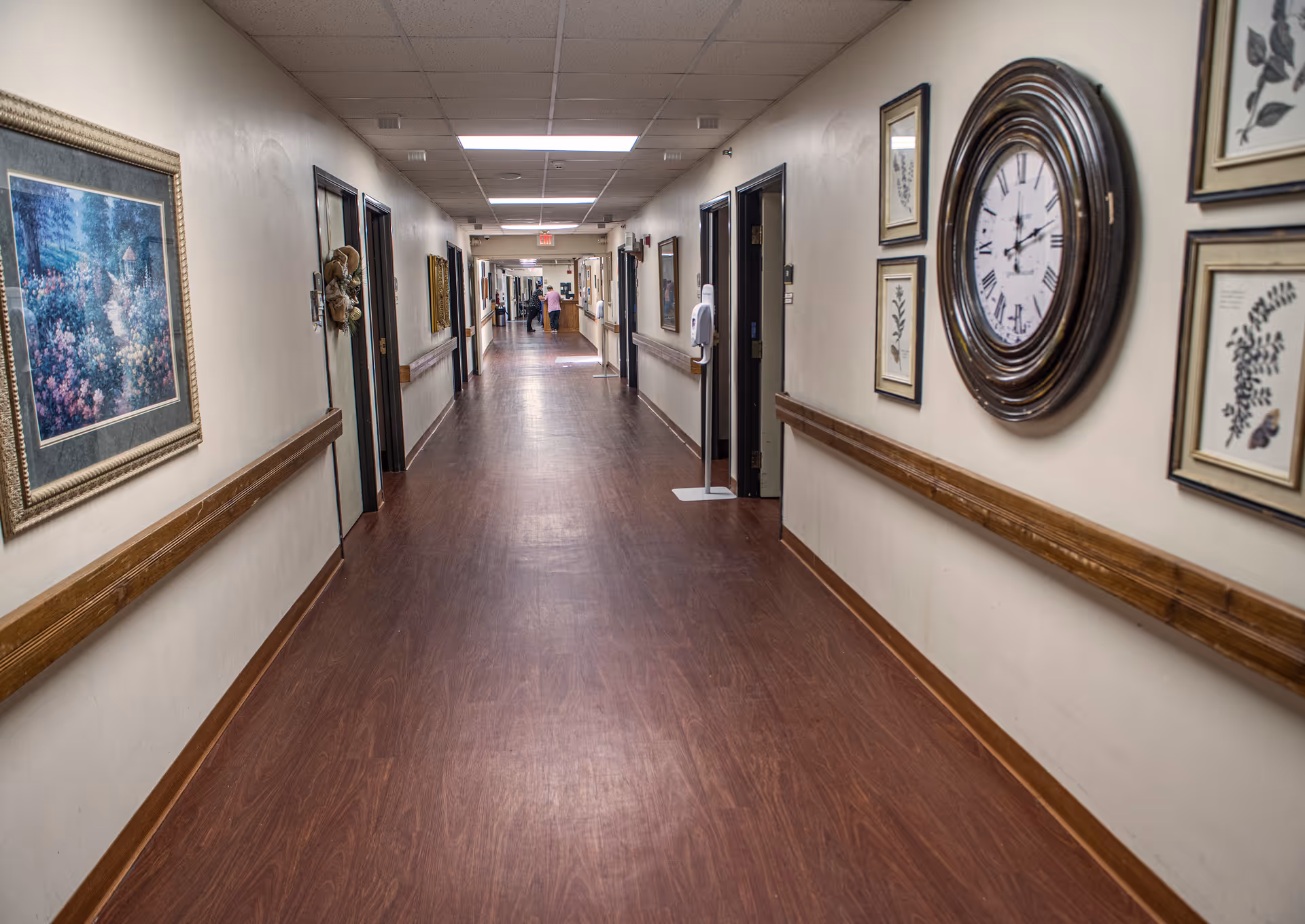 A long hallway in a senior living facility with wooden floors and handrails on both sides. The walls are decorated with framed artwork and a large round clock. Several open doorways line the hallway, and a person is visible in the distance near the end of the corridor.