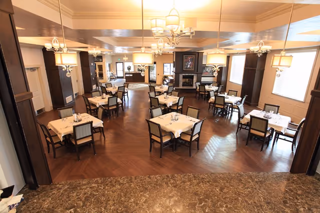 A spacious dining room in an assisted living facility with multiple square tables covered with white tablecloths, each surrounded by four chairs. The room features wooden flooring, elegant chandeliers, large windows with blinds, and a fireplace with artwork above it in the background.