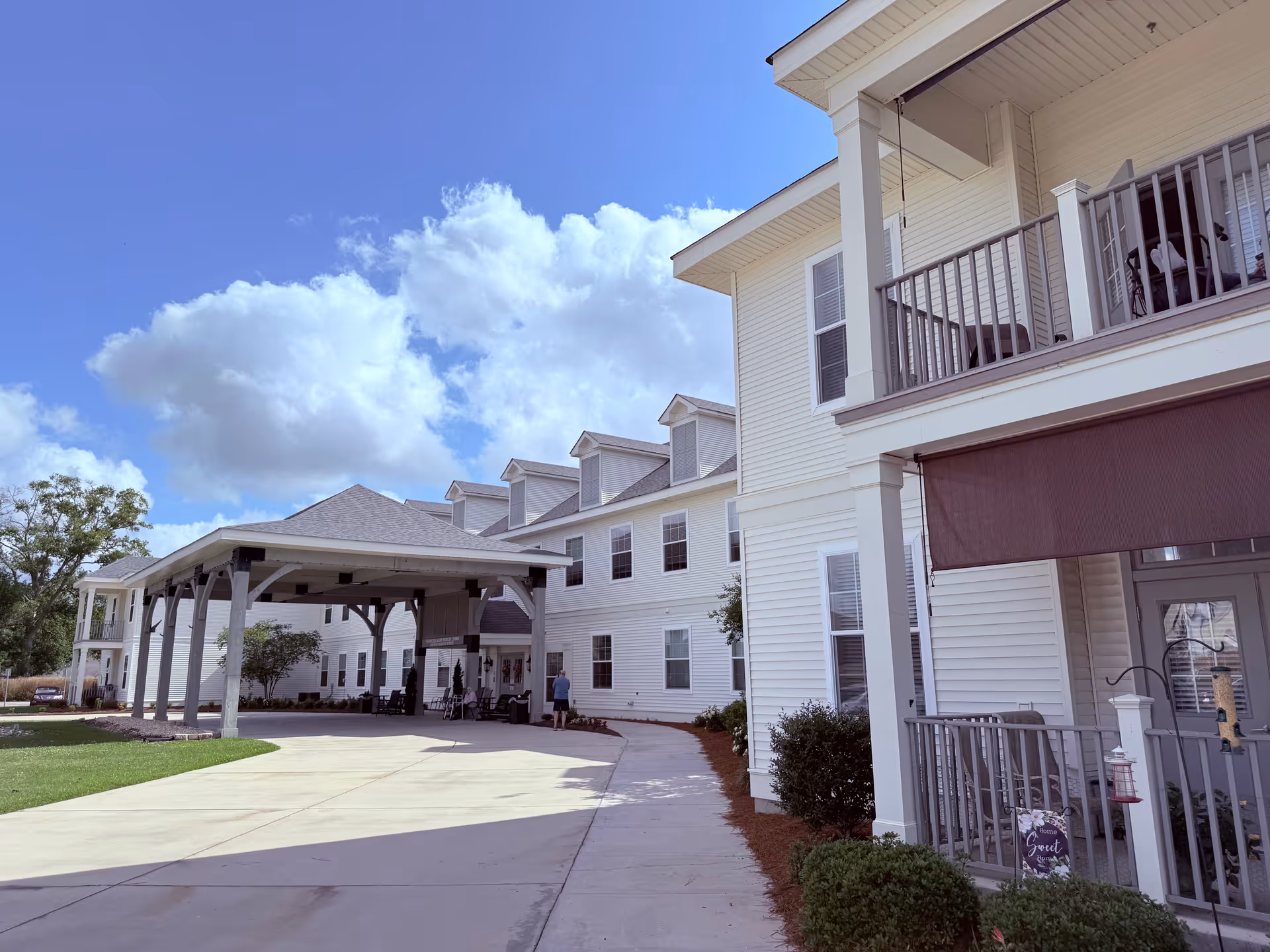 Exterior view of a senior living facility with white siding and multiple windows. The building has a covered entrance with a driveway and a sidewalk leading to the entrance. There are balconies with railings and some outdoor furniture. The sky is blue with scattered clouds.
