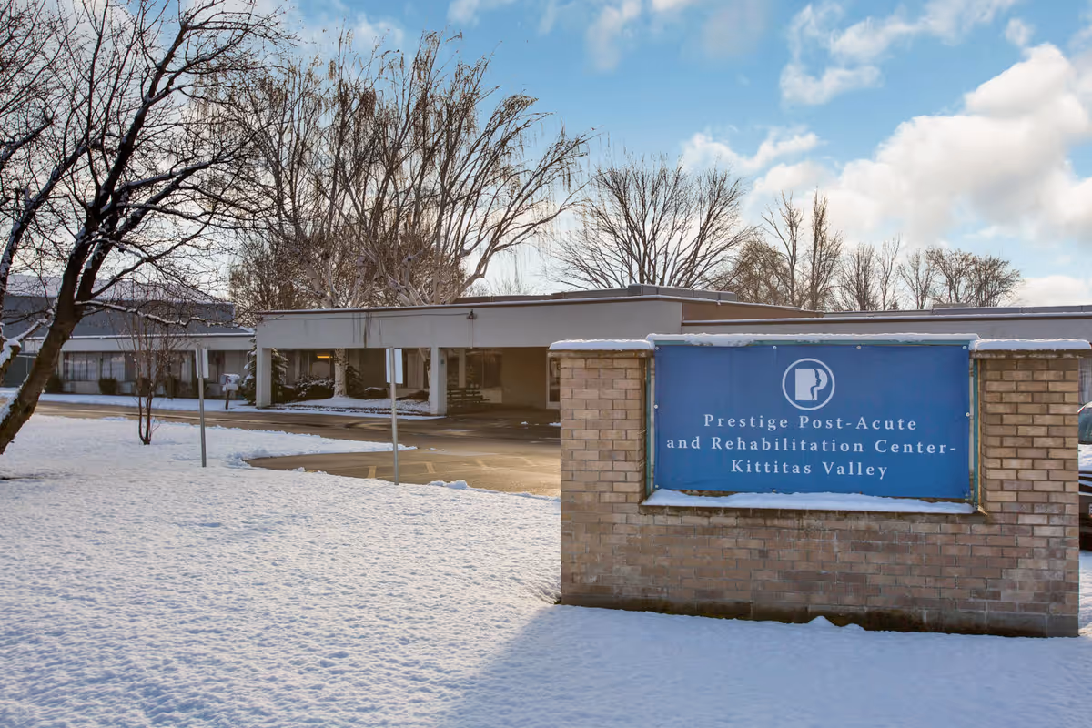 Snow-covered entrance of Prestige Post-Acute & Rehabilitation Center - Kittitas Valley with a brick sign in the foreground and the one-story building and trees behind it.