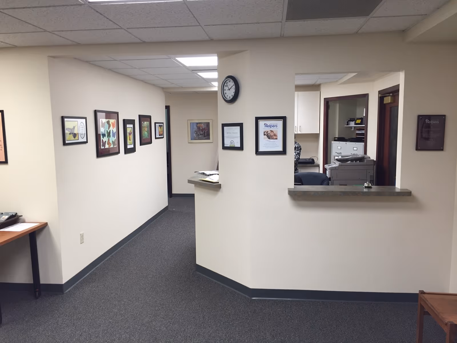 Interior hallway of a senior facility with wall art, a clock, and a reception window.