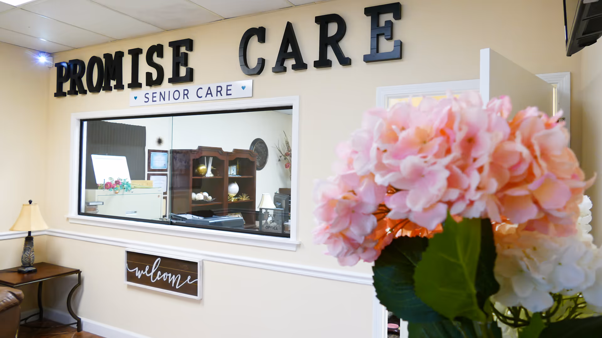 Interior reception area of Promise Care Senior Care with a windowed reception desk and pink flowers