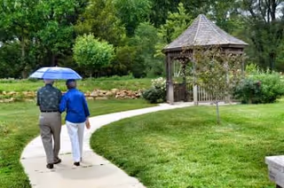 Two elderly people walk along a curved path with a blue umbrella toward a gazebo.