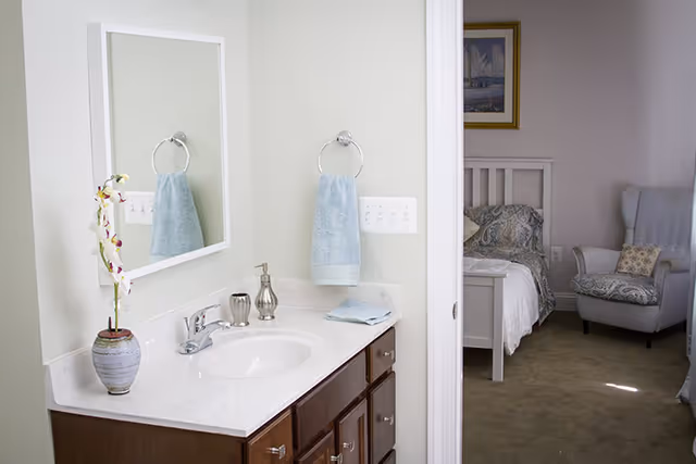 Bathroom vanity with sink, mirror, towels, and doorway to a bedroom in Cobbdale Assisted Living.